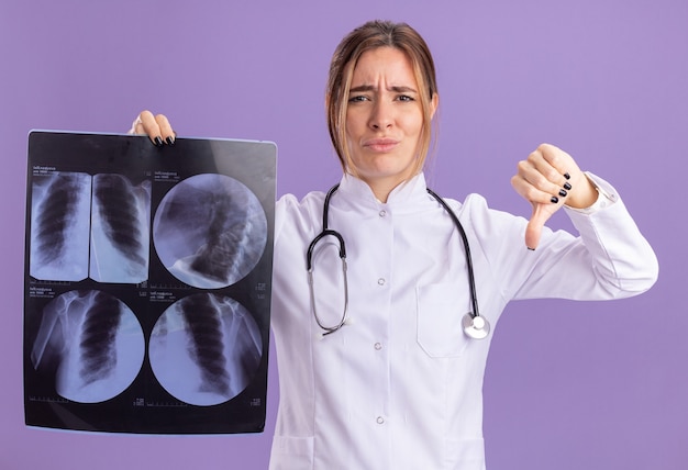 Jeune femme médecin mécontente portant une robe médicale avec stéthoscope tenant une radiographie montrant le pouce vers le bas isolé sur un mur violet