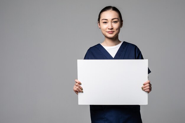 Jeune femme médecin montrant un panneau d'affichage vierge, isolé sur un mur blanc