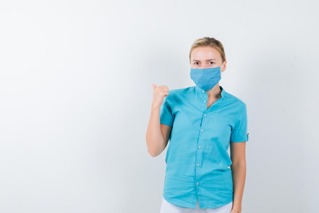 Jeune femme médecin pointant vers l'arrière avec le pouce en uniforme médical