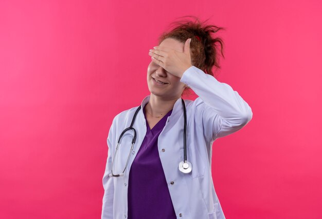 Jeune femme médecin portant blouse blanche avec stéthoscope couvrant les yeux avec bras souriant debout sur un mur rose