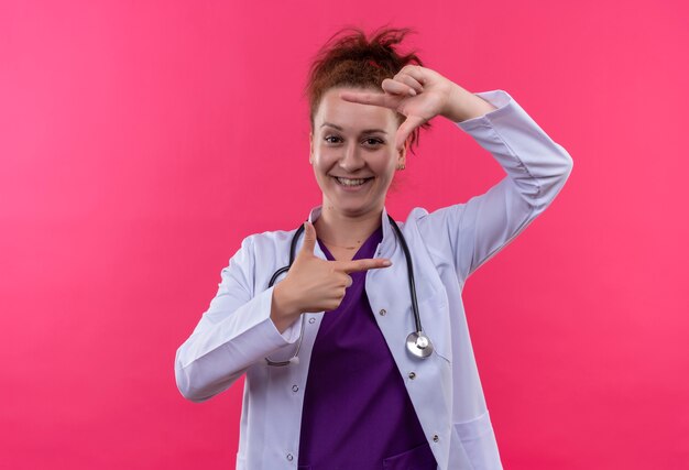 Jeune femme médecin portant une blouse blanche avec stéthoscope faisant cadre avec les doigts souriant joyeusement regardant à travers ce cadre debout sur un mur rose