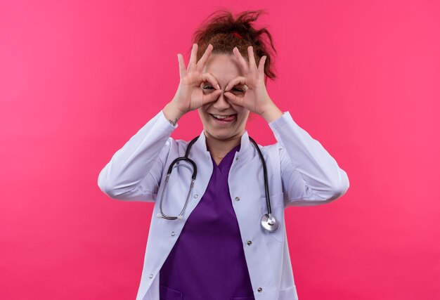 Jeune femme médecin portant blouse blanche avec stéthoscope faisant signe ok avec les doigts comme des jumelles regardant à travers les doigts qui sort la langue debout sur le mur rose