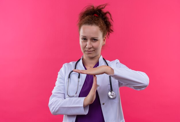 Jeune femme médecin portant une blouse blanche avec stéthoscope à la fatigue et l'ennui faisant le geste de temps avec les mains debout sur rose