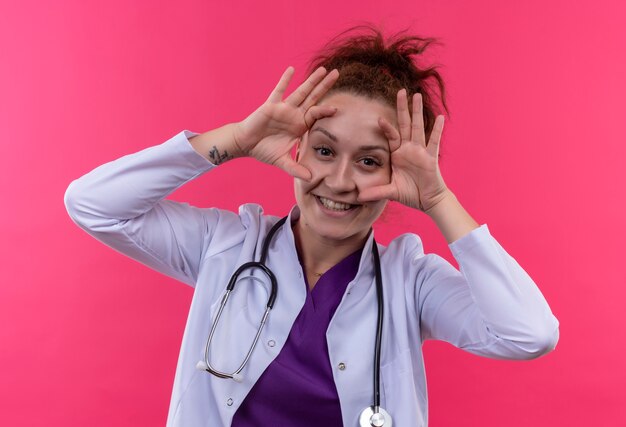 Jeune femme médecin portant une blouse blanche avec stéthoscope ouvrant les yeux avec les doigts surpris et heureux debout sur le mur rose