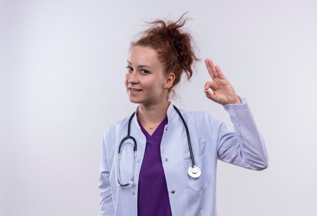 Jeune femme médecin portant blouse blanche avec stéthoscope souriant confiant faisant signe ok debout sur un mur blanc