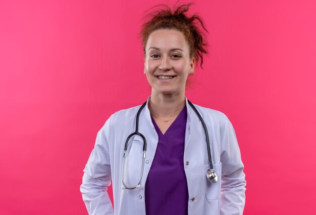 Jeune femme médecin portant blouse blanche avec stéthoscope souriant largement debout sur un mur rose