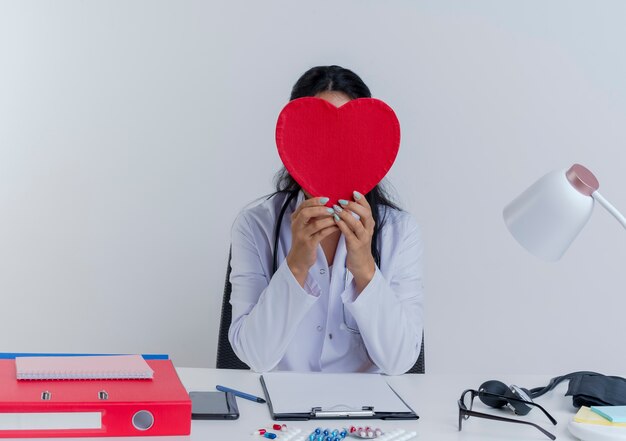 Jeune femme médecin portant une robe médicale et un stéthoscope assis au bureau avec des outils médicaux tenant et se cachant derrière la forme de coeur isolé