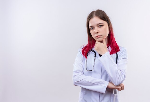 Jeune femme médecin portant une robe médicale stéthoscope mettant la main sur le menton sur un mur blanc isolé avec copie espace