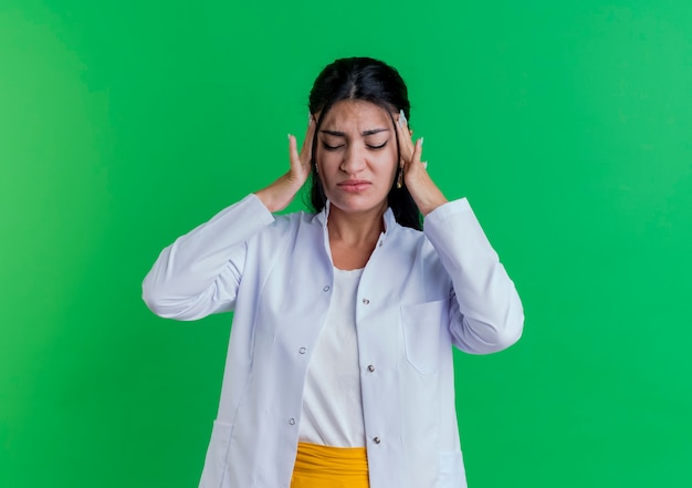 Jeune femme médecin portant une robe médicale tenant la tête souffrant de maux de tête avec les yeux fermés isolé sur un mur vert avec espace copie