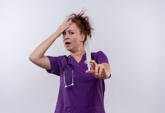 Jeune femme médecin portant l'uniforme médical avec stéthoscope tenant la seringue peur et inquiet debout sur un mur blanc