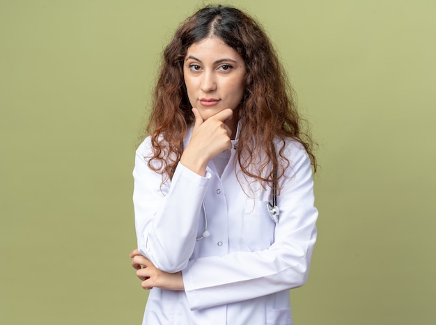 Jeune Femme Médecin Réfléchie Portant Une Robe Médicale Et Un Stéthoscope Gardant La Main Sur Le Menton Isolée Sur Un Mur Vert Olive Avec Espace De Copie