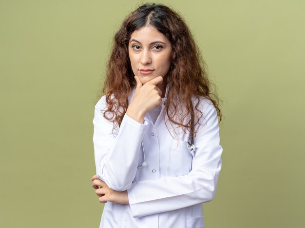 Jeune femme médecin réfléchie portant une robe médicale et un stéthoscope gardant la main sur le menton isolée sur un mur vert olive avec espace de copie