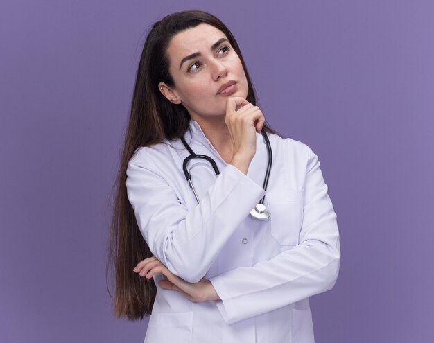 Une jeune femme médecin réfléchie portant une robe médicale avec un stéthoscope tient le menton en levant