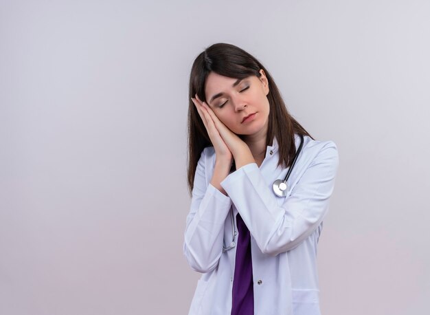 Jeune femme médecin en robe médicale avec stéthoscope fait semblant de dormir les yeux fermés sur un mur blanc isolé