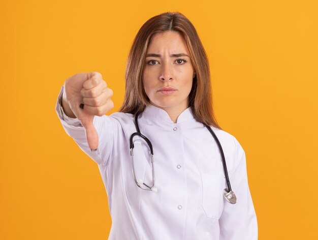 Jeune femme médecin stricte portant une robe médicale avec stéthoscope montrant le pouce vers le bas isolé sur mur jaune