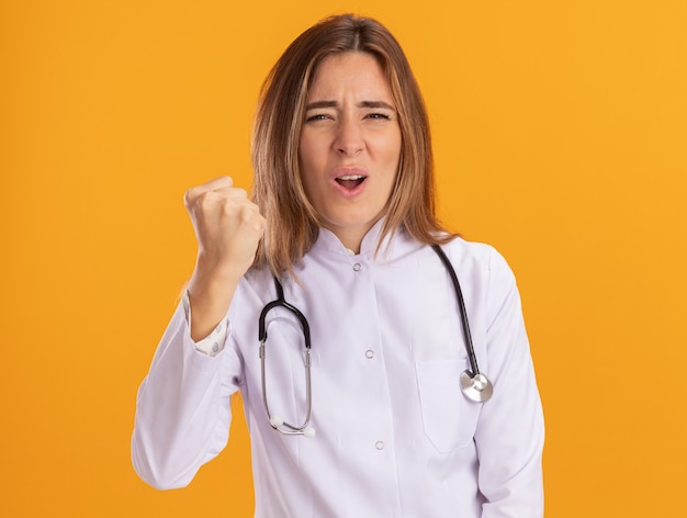 Jeune femme médecin stricte portant une robe médicale avec stéthoscope tenant le poing isolé sur mur jaune