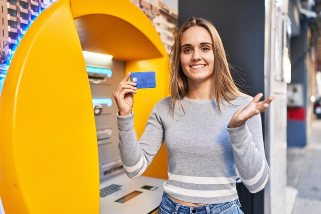 Jeune femme médecin tenant une carte de crédit au distributeur de billets célébrant la réussite avec un sourire heureux et une expression gagnante avec la main levée