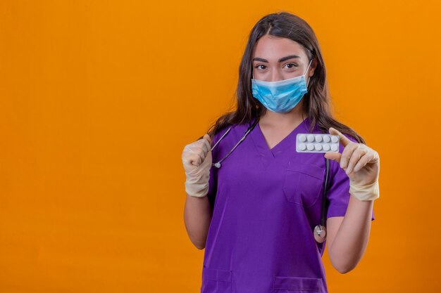 Jeune femme médecin en uniforme médical avec phonendoscope portant un masque de protection et des gants smiling holding blister avec des pilules et le poing levé sur fond orange isolé