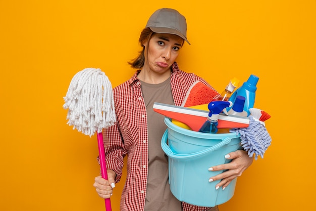 Jeune femme de ménage bouleversée en chemise à carreaux et casquette tenant un seau avec des outils de nettoyage et une vadrouille regardant la caméra avec une expression triste pinçant les lèvres debout sur fond orange