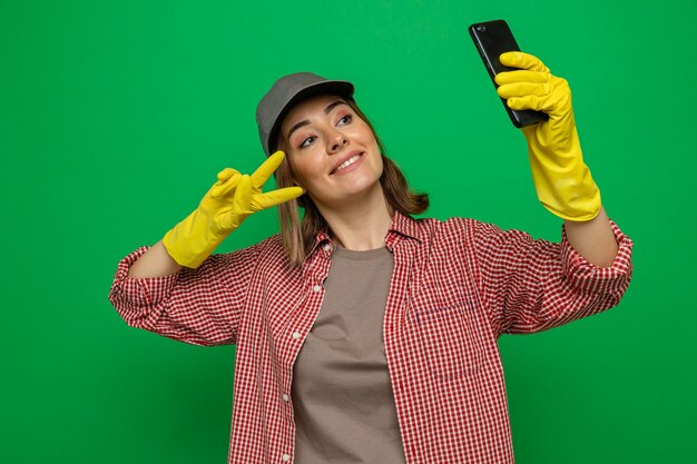 Jeune femme de ménage en chemise à carreaux et casquette portant des gants en caoutchouc faisant du selfie à l'aide d'un smartphone souriant joyeusement montrant le signe v