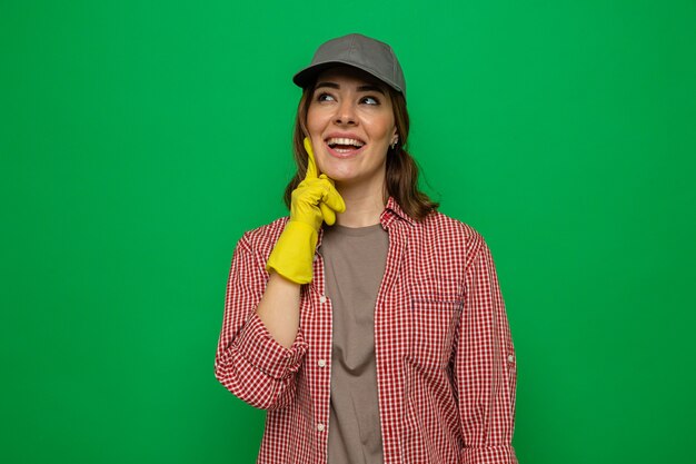 Jeune femme de ménage en chemise à carreaux et casquette portant des gants en caoutchouc levant souriant joyeusement rêvant debout sur fond vert