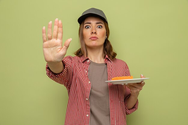 Jeune femme de ménage en chemise à carreaux et casquette tenant une assiette et une éponge regardant la caméra avec un visage sérieux faisant un geste d'arrêt avec la main debout sur fond vert