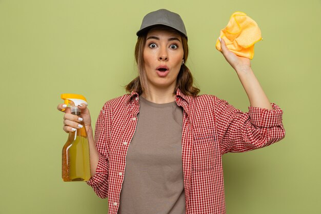 Jeune femme de ménage en chemise à carreaux et casquette tenant un chiffon et un spray de nettoyage regardant la caméra surpris debout sur fond vert