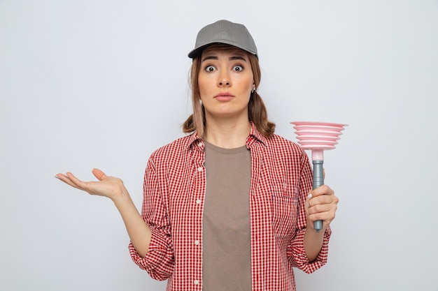 Jeune femme de ménage en chemise à carreaux et casquette tenant le piston regardant la caméra surpris et confus debout sur fond blanc
