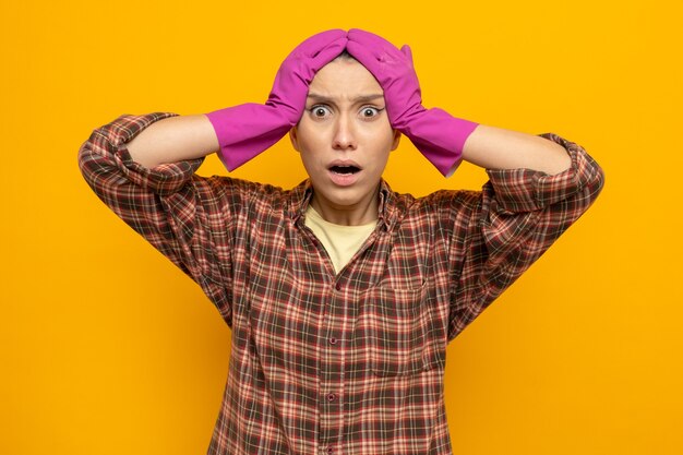 Jeune femme de ménage en chemise à carreaux dans des gants en caoutchouc confuse et inquiète se tenant la main sur la tête pour erreur debout sur orange