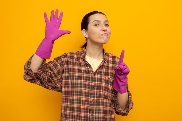 Jeune femme de ménage en chemise à carreaux dans des gants en caoutchouc avec un sourire confiant sur le visage montrant le numéro six avec les doigts debout sur l'orange