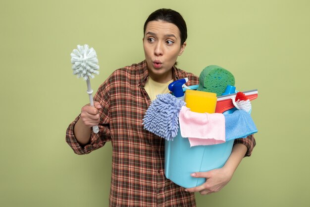 Jeune femme de ménage en chemise à carreaux tenant une brosse de nettoyage et un seau avec des outils de nettoyage regardant une brosse confuse debout sur le vert