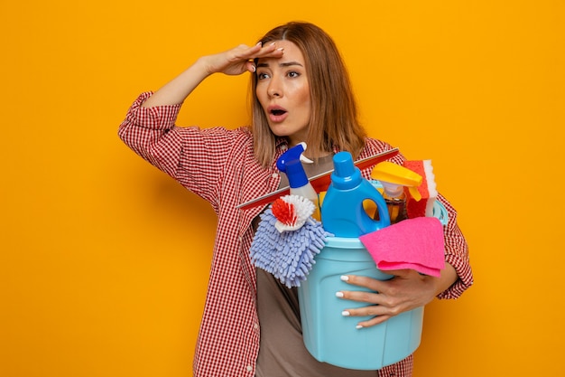 Jeune femme de ménage en chemise à carreaux tenant un seau avec des outils de nettoyage regardant au loin avec la main sur la tête pour regarder quelque chose ou quelqu'un