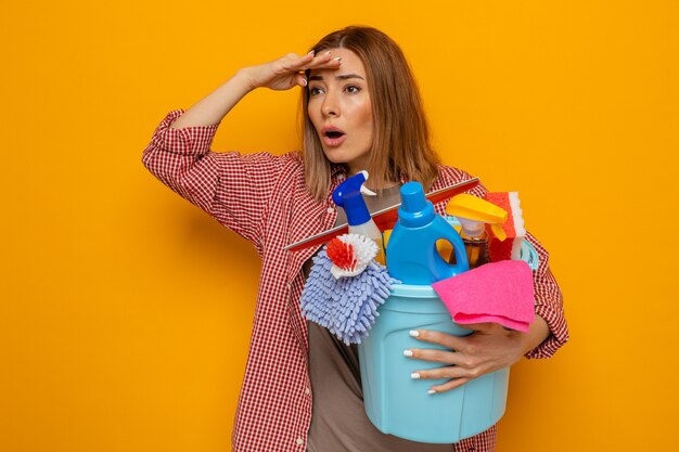 Jeune femme de ménage en chemise à carreaux tenant un seau avec des outils de nettoyage regardant au loin avec la main sur la tête pour regarder quelque chose ou quelqu'un