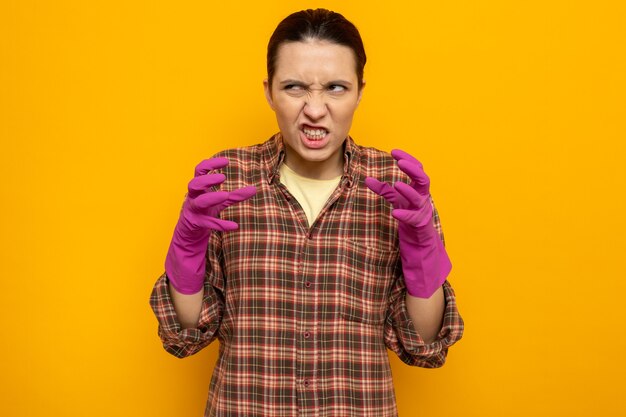 Jeune femme de ménage dans des vêtements décontractés dans des gants en caoutchouc faisant la bouche tordue en colère et frustrée par les bras levés debout sur l'orange