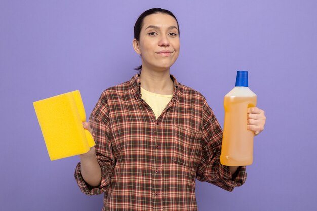 Jeune femme de ménage dans des vêtements décontractés tenant une bouteille de produits de nettoyage avec une éponge regardant à l'avant un sourire heureux et positif confiant sur le mur violet