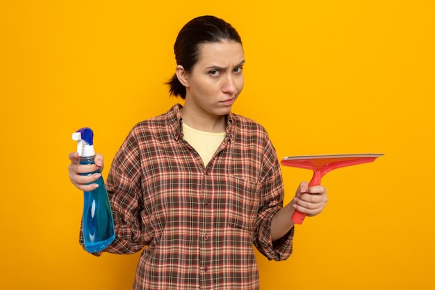Jeune femme de ménage dans des vêtements décontractés tenant un spray de nettoyage et une vadrouille à la recherche d'un visage fronçant les sourcils