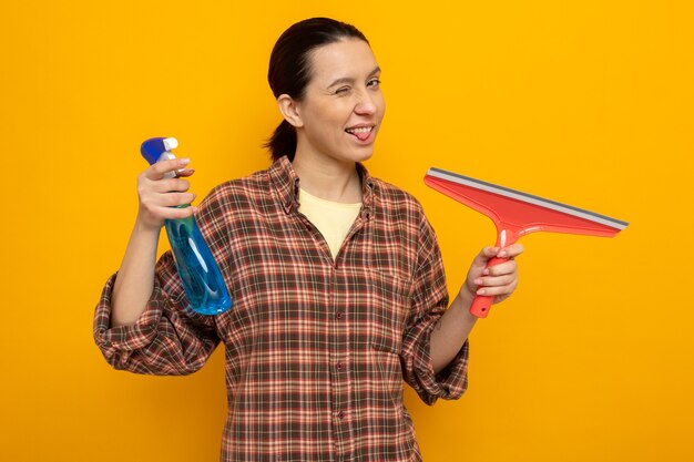 Jeune femme de ménage dans des vêtements décontractés tenant un spray de nettoyage et une vadrouille regardant l'avant heureux et positif qui sort la langue debout sur le mur orange