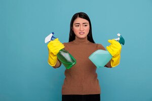 Jeune femme de ménage terne inactive asiatique des corvées tenant deux sprays détergents dans des gants jaunes sur fond bleu, concept de maison de nettoyage, mécontent étonné d'un état d'esprit négatif,