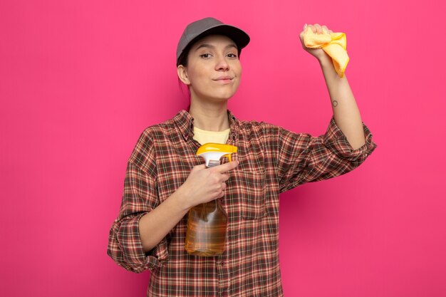 Jeune femme de ménage en vêtements décontractés et casquette tenant un chiffon et un spray de nettoyage souriant confiant prêt pour le nettoyage debout sur rose