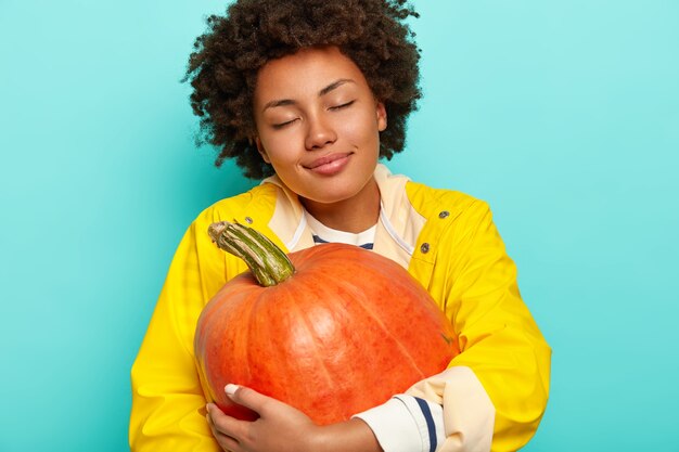 Une jeune femme métisse satisfaite tient la citrouille récoltée orange, porte un imperméable jaune décontracté, a les yeux fermés, pose sur fond bleu.