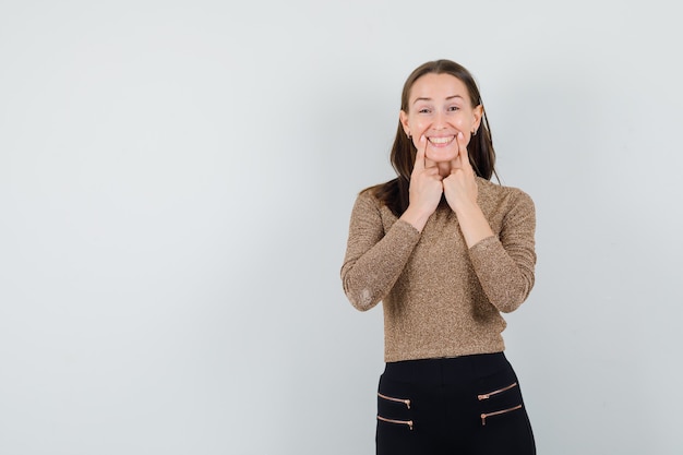 Jeune femme mettant l'index près de la bouche, forçant le sourire en pull doré et pantalon noir et à la recherche de bonheur. vue de face.