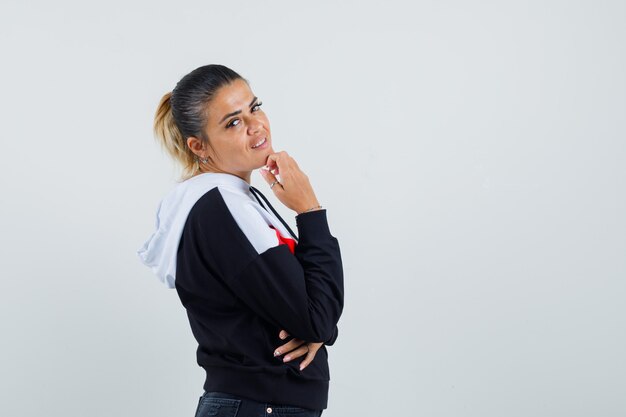 Jeune femme mettant la main sur le menton, pensant à quelque chose en pull et jeans noirs et à la pensif. vue de face.