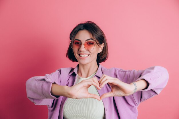 Jeune femme mignonne vêtements décontractés lunettes de soleil sur fond rose heureux actif montrant coeur avec les mains