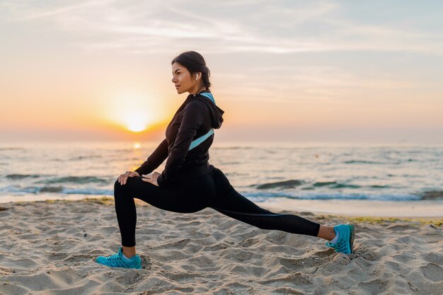 Jeune femme mince attrayante, faire des exercices de sport sur la plage du lever du soleil du matin en vêtements de sport, mode de vie sain, écouter de la musique sur les écouteurs, faire des étirements pour les jambes