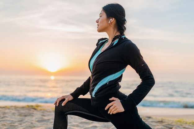 Jeune femme mince attrayante, faire des exercices de sport sur la plage du lever du soleil du matin en vêtements de sport, mode de vie sain, écouter de la musique sur les écouteurs, faire des étirements