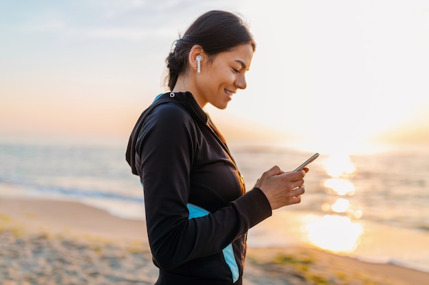 Jeune femme mince attrayante, faire des exercices de sport sur la plage du lever du soleil du matin en vêtements de sport, mode de vie sain, écouter de la musique sur des écouteurs sans fil tenant un smartphone, souriant heureux