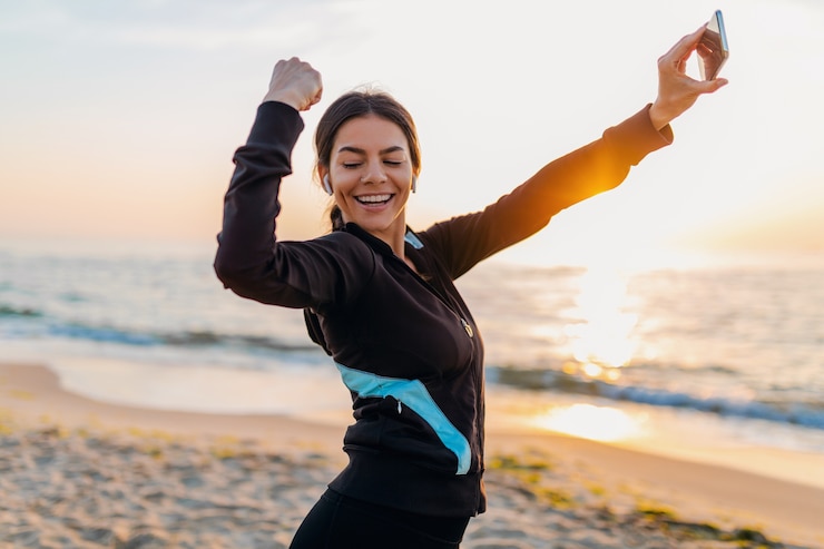 Photo gratuit jeune femme mince attrayante souriante, faire des exercices de sport sur la plage du lever du soleil du matin dans les vêtements de sport, mode de vie sain, écouter de la musique sur les écouteurs, faire selfie photo sur téléphone à la recherche forte