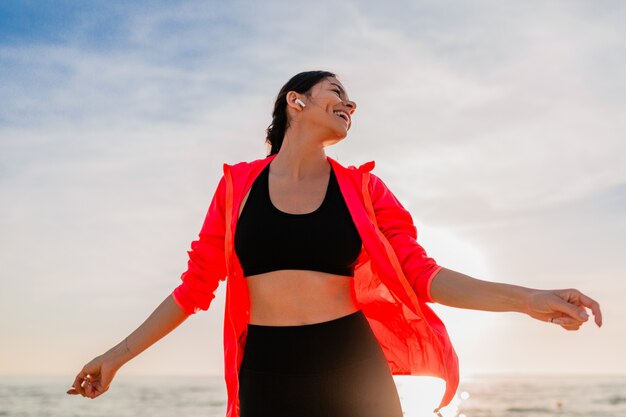 Jeune femme mince attrayante souriante faisant du sport au lever du soleil du matin dansant sur la plage de la mer en vêtements de sport, mode de vie sain, écouter de la musique sur les écouteurs, porter une veste coupe-vent rose, s'amuser