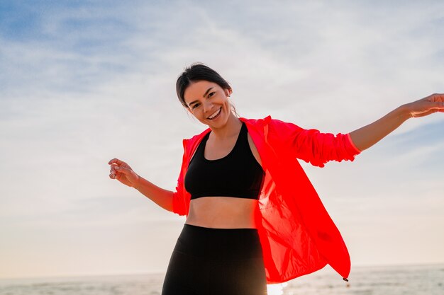 Jeune femme mince attrayante souriante faisant du sport au lever du soleil du matin dansant sur la plage de la mer en vêtements de sport, mode de vie sain, écouter de la musique sur les écouteurs, porter une veste coupe-vent rose, s'amuser