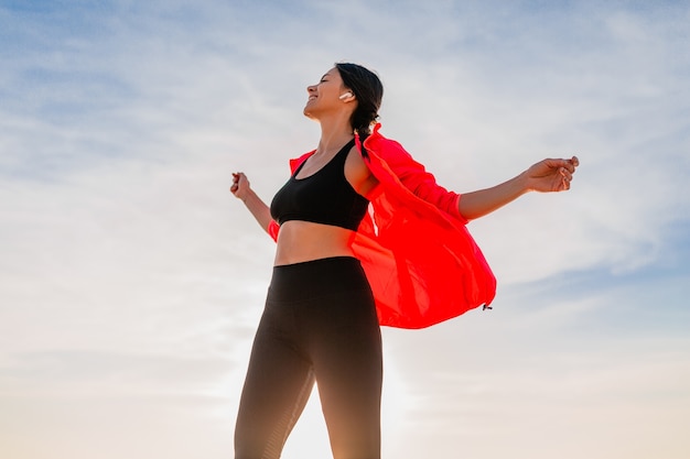 Jeune femme mince attrayante souriante faisant du sport au lever du soleil du matin dansant sur la plage de la mer en vêtements de sport, mode de vie sain, écouter de la musique sur les écouteurs, porter une veste coupe-vent rose, s'amuser